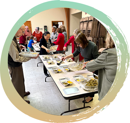 Two large tables pictured, both having foil food containers with the lids off lined up on them. Many people holding different food items are walking around the table placing their item into each container.