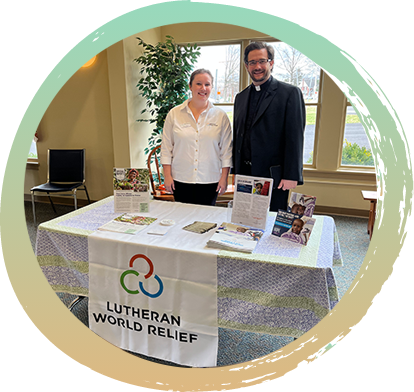 Pastor Matt and a lady wearing a white shirt and black pants are standing behind a table. One the table there are several pamphlets. In front of the table is a sign that reads "Lutheran World Relief" on it.