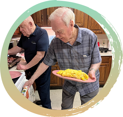 Two men standing in the kitchen cooking. One man, with a dark blue shirt is at the counter cooking at a skillet. The other man has cooked eggs on a pink plate and he is reaching for the oven door.
