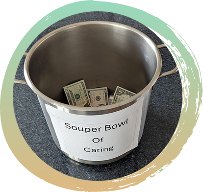 A metal pail with a sign that reads "Souper Bowl Of Caring" taped to the outside, with money laying inside the pail.