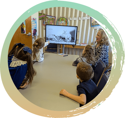 A woman teaching a Sunday School lesson, is sitting at a table with four children. At the end of the table is a screen with a black and white image of a horse and a donkey grazing in a field.