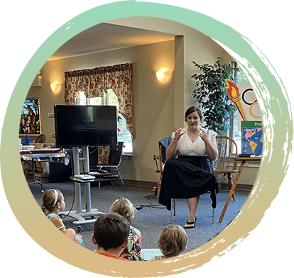 A woman wearing a white shirt and black skirt is sitting on a chair in front of a group of children teaching a Vacation Bible School Lesson. There is a sign behind her that has the olympic rings and torch that reads "The World" on a easel. VBS Lesson 8’12’24