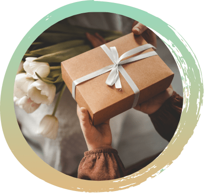 Someone handing a brown box with a white ribbon to someone else. In the background is a white backdrop with a white bunch of roses